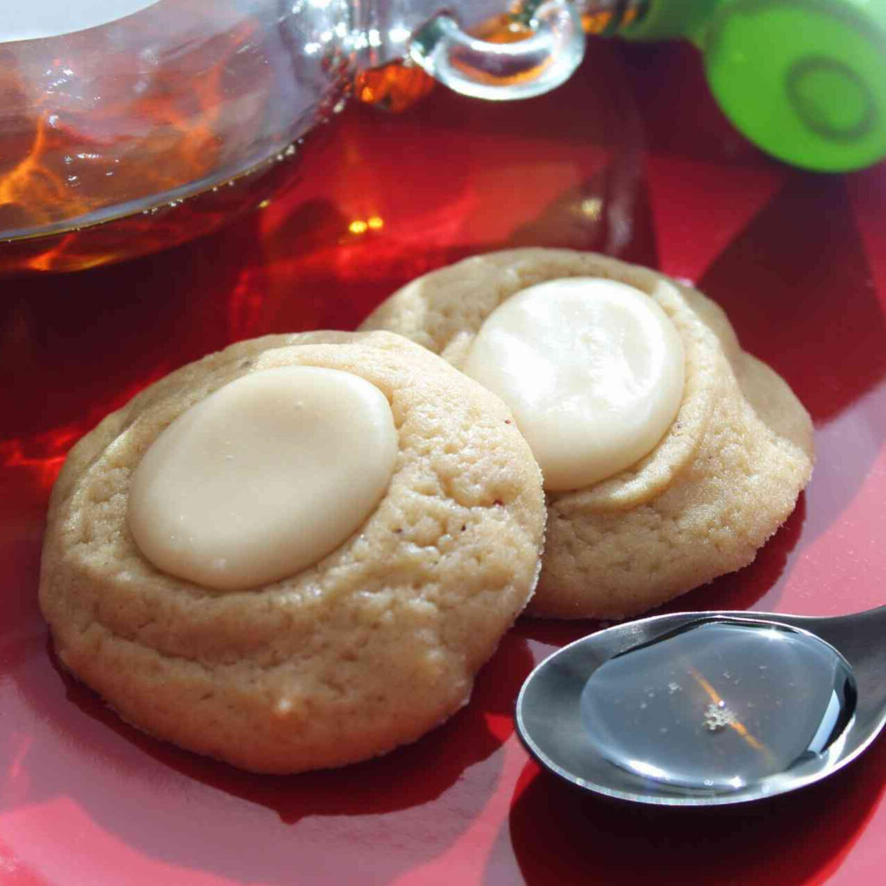 A photo of two thumbprint cookies on a red plate, with a spoon full of syrup in front and a cropped syrup bottle behind.
